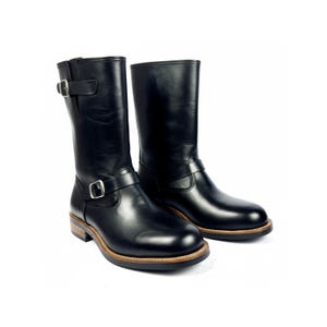 Puede incluir: Un par de botas de moto de cuero negro. Las botas son de media caña con correas de hebilla. Las suelas tienen un borde marrón y una suela marrón claro. La altura de las botas es de aproximadamente 25 cm.