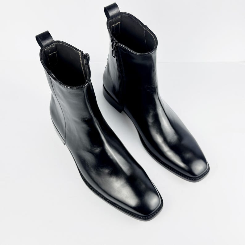 May include: A pair of black leather ankle boots with a square toe and a side zip. The boots have a pull tab at the back and a low heel. The leather has a glossy finish.