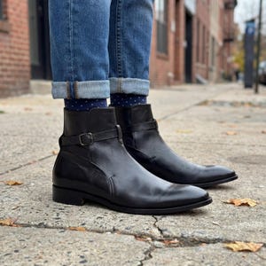 May include: A pair of black leather ankle boots with a buckle strap detail. The boots have a low heel and are worn with blue jeans and patterned socks. The boots are shown outdoors on a concrete surface.