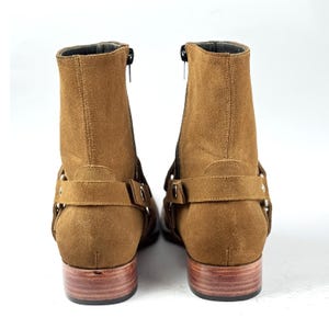 May include: A pair of brown suede ankle boots with side zippers and decorative straps. The boots have a low heel and a wooden sole. The boots are shown against a white background.