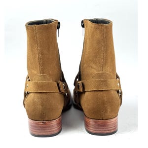 May include: A pair of brown suede ankle boots with side zippers and decorative straps. The boots have a low wooden heel and a rounded toe. The boots are shown against a white background.