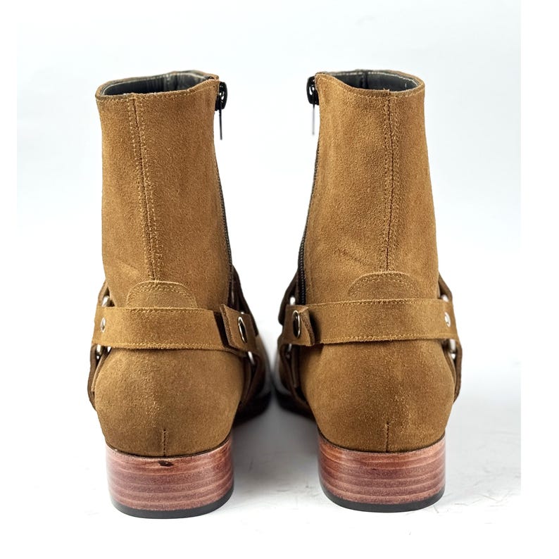 May include: A pair of brown suede ankle boots with side zippers and decorative straps. The boots have a low wooden heel and a rounded toe. The boots are shown against a white background.