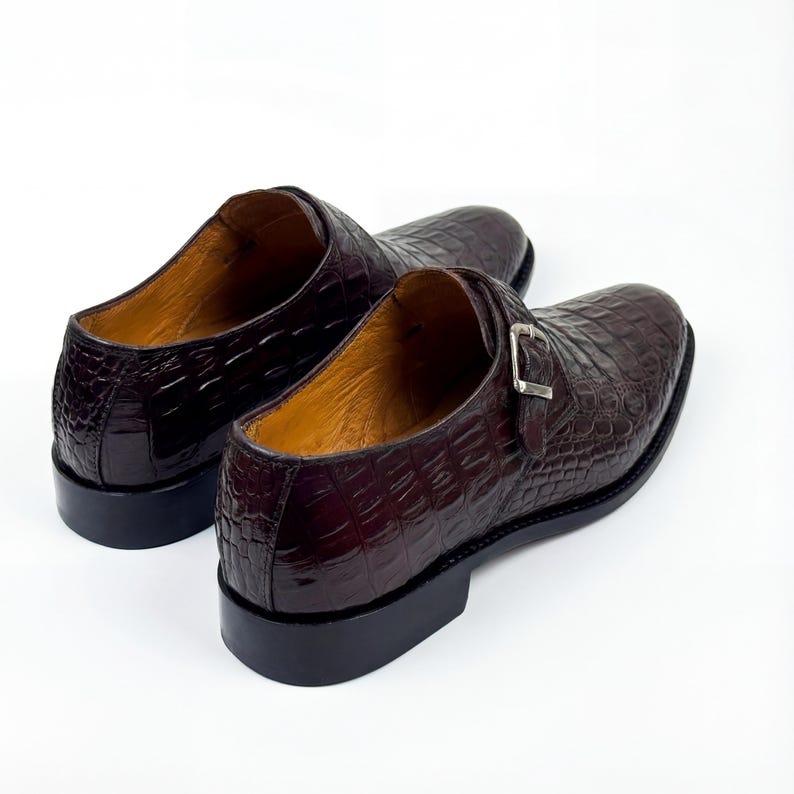 May include: A pair of dark brown leather dress shoes with a crocodile skin texture. The shoes have a buckle closure and a black sole. The interior of the shoes is a light brown color.