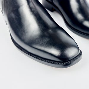 May include: Close-up of a pair of black leather boots. The boots have a square toe and a smooth, shiny finish. The stitching around the sole is visible. The boots are on a white background.