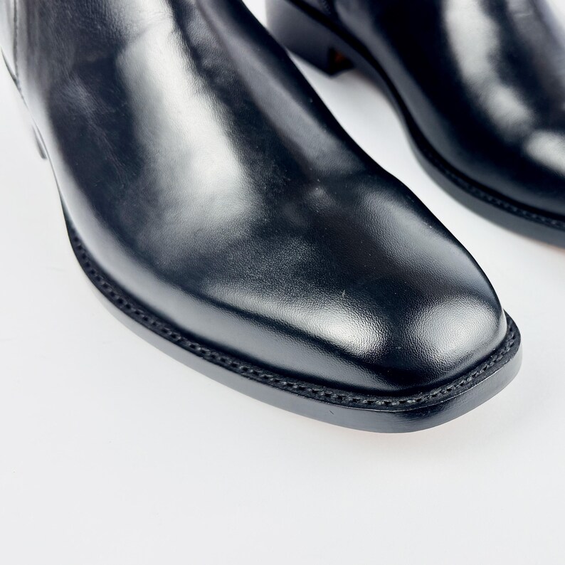 May include: Close-up of a pair of black leather boots. The boots have a square toe and a smooth, shiny finish. The stitching around the sole is visible. The boots are on a white background.