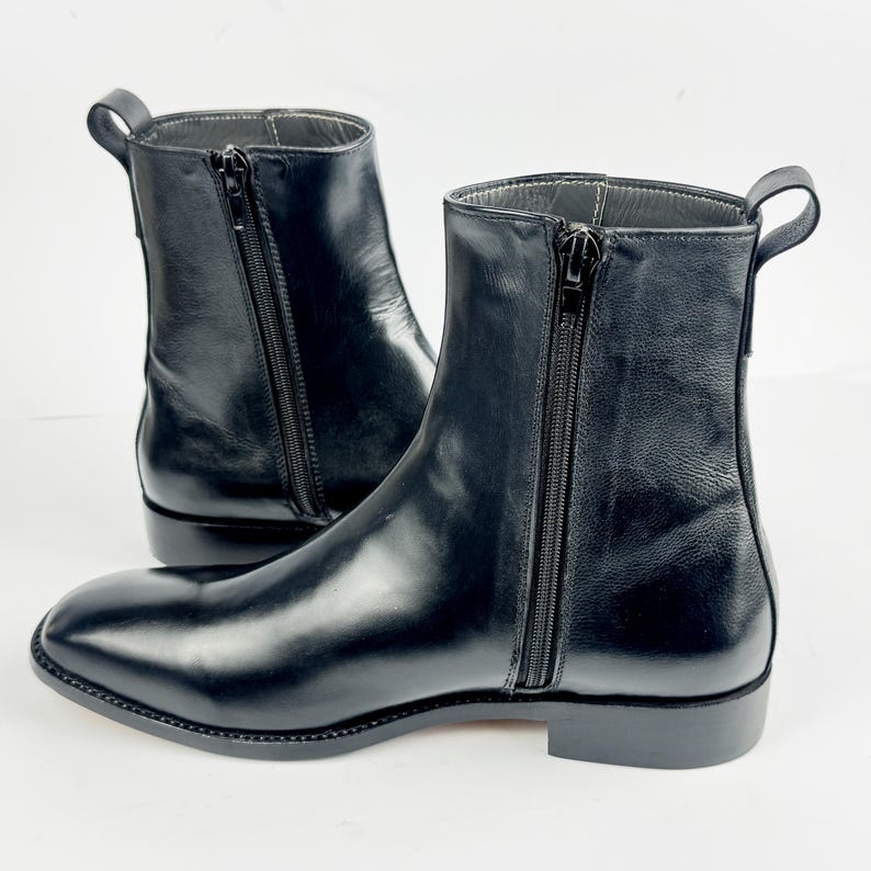 May include: A pair of black leather ankle boots. The boots feature a side zip, a small heel, and a pull tab. The leather has a glossy finish. The boots are set against a white background.