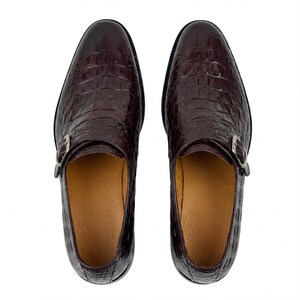 May include: A pair of dark brown leather dress shoes with a crocodile skin texture. The shoes have a single silver buckle closure and a rounded toe. The interior is a light tan color.