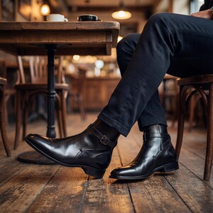 May include: A person seated in a cafe wearing black leather ankle boots with a buckle closure. The boots have a polished finish and are paired with black trousers. The setting includes a wooden table and chairs.