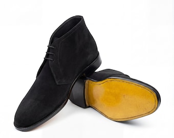 May include: A pair of black suede chukka boots. The boots have a lace-up closure and a rounded toe. The soles are a light yellow color. The boots are set against a white background.