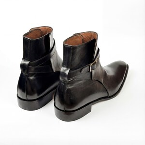 May include: A pair of black leather ankle boots with a square toe and a strap with a gold-colored buckle. The boots have a glossy finish and a brown interior. The boots are displayed on a white background.