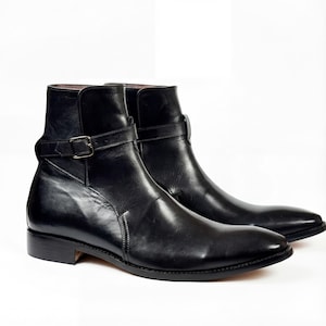 May include: A pair of black leather ankle boots with a pointed toe and a strap with a buckle detail. The boots have a low heel and a smooth, polished finish. The boots are displayed on a white background.