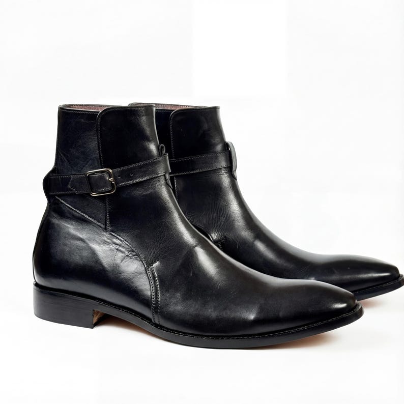 May include: A pair of black leather ankle boots with a pointed toe and a strap with a buckle detail. The boots have a low heel and a smooth, polished finish. The boots are displayed on a white background.