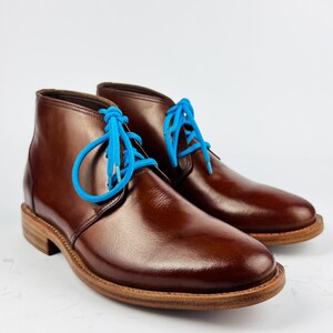May include: A pair of brown leather chukka boots with bright blue laces. The boots have a polished finish and a light brown sole. The laces are a vibrant contrast to the dark brown leather. The boots are displayed on a white background.