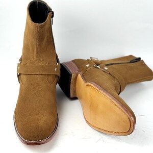 May include: A pair of brown suede ankle boots with a side zipper and a decorative strap with silver-tone hardware. The boots have a stacked heel and a leather sole. The boots are approximately 6 inches tall.