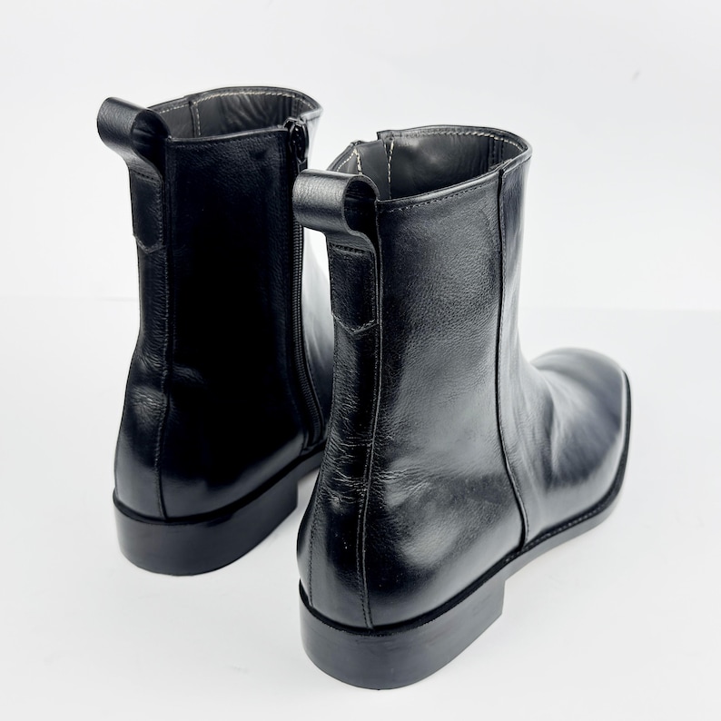 May include: A pair of black leather ankle boots with a square toe and a low heel. The boots have a side zip and a pull tab. The boots are approximately 15 cm tall.