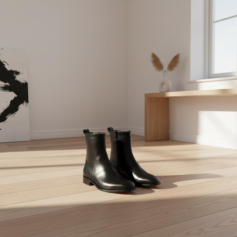 May include: A pair of black leather ankle boots with a low heel. The boots have a sleek, minimalist design with a zipper on the side. They are placed on a light-coloured wooden floor, with natural light illuminating the scene.