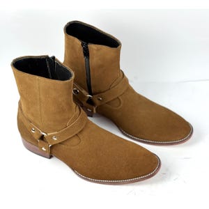 May include: A pair of brown suede ankle boots. The boots feature a side zipper, a strap with a metal buckle, and a stitched sole. The boots have a low heel and a rounded toe.