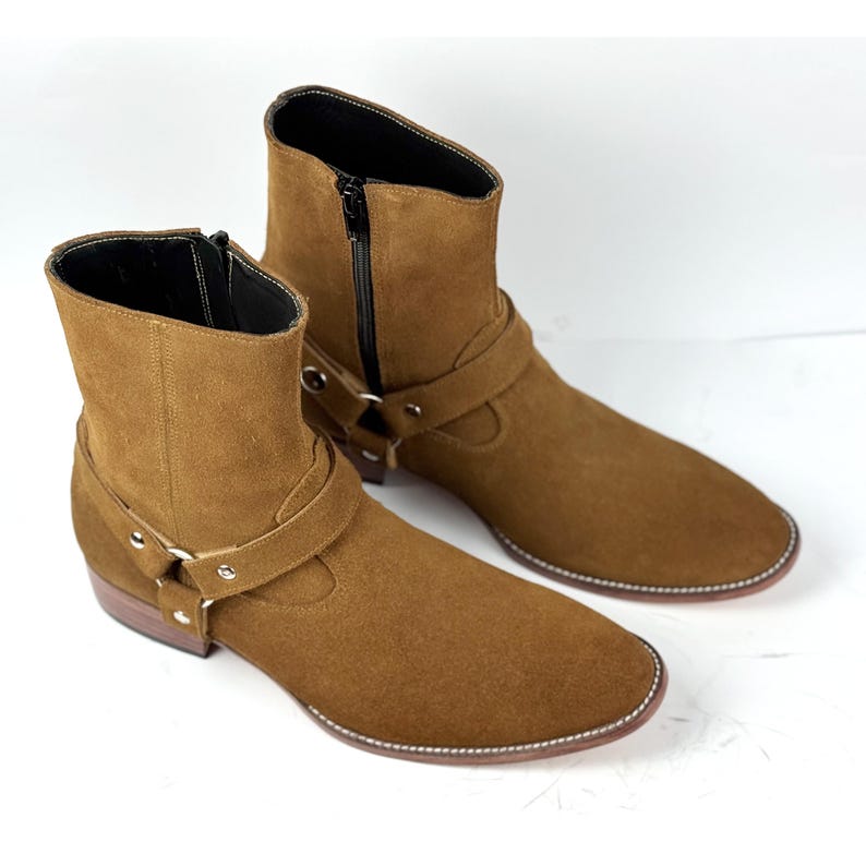 May include: A pair of brown suede ankle boots. The boots feature a side zipper, a strap with a metal buckle, and a stitched sole. The boots have a low heel and a rounded toe.