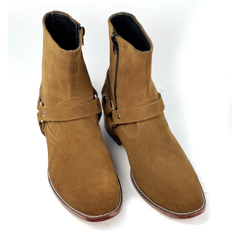 May include: A pair of brown suede ankle boots with a side zipper and a strap with a buckle. The boots have a rounded toe and a stitched sole. The boots are displayed on a white background.
