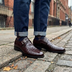 May include: A pair of dark brown leather monk strap shoes with a crocodile texture. The shoes have a silver buckle and are worn with blue jeans and patterned socks. The shoes are on a cobblestone street.