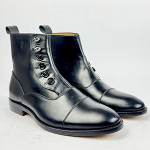 Puede incluir: Botines de cuero negro con diseño de puntera. Los botines tienen cierre de botones y una lengüeta. Los botines tienen un tacón bajo y un acabado liso. Miden aproximadamente 15 cm de alto.