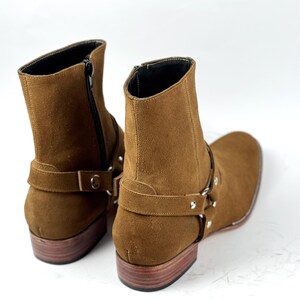 May include: Brown suede ankle boots with a square toe and a low wooden heel. The boots have a side zipper and a strap with silver-tone hardware. The boots are a neutral color and are suitable for various outfits.