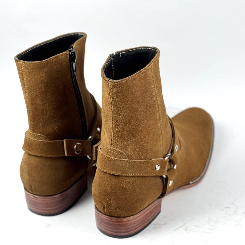 May include: Brown suede ankle boots with a square toe and a low wooden heel. The boots have a side zipper and a strap with silver-tone hardware. The boots are a neutral color and are suitable for various outfits.