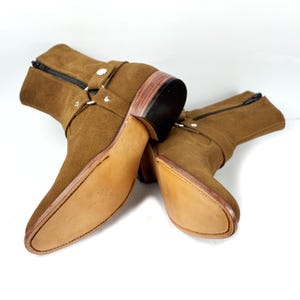 May include: A pair of brown suede ankle boots with a pointed toe and a stacked heel. The boots have a side zipper and a strap with silver-tone hardware. The soles are a light brown color.