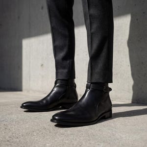 May include: Black leather ankle boots with a buckle detail. The boots are worn with dark gray trousers. The boots have a sleek, polished appearance and a low heel. The image is taken from a low angle, highlighting the footwear.