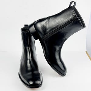 May include: A pair of black leather ankle boots with a polished finish. The boots have a side zip, a pull tab, and a low heel. The design is minimalist and modern, suitable for various outfits. The boots are approximately 15 cm high.