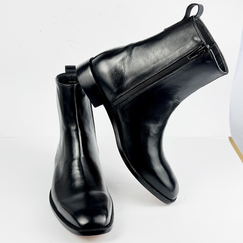 May include: A pair of black leather ankle boots with a polished finish. The boots have a side zip, a pull tab, and a low heel. The design is minimalist and modern, suitable for various outfits. The boots are approximately 15 cm high.