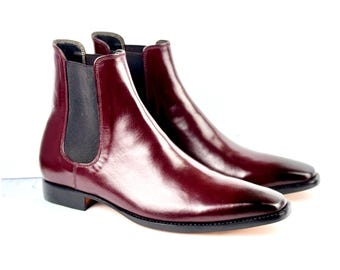 May include: A pair of burgundy leather Chelsea boots. The boots have a polished, glossy finish and feature elastic side panels in black. The soles are black, and the boots are set against a white background.