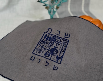Könnte beinhalten: Eine graue Stoffabdeckung mit dunkelblauer Stickerei eines quadratischen Rahmens, der botanische Illustrationen von Weizen, Trauben und einem Granatapfel enthält. Der hebräische Text "שבת שלום" ist dunkelblau über und unter dem Design gestickt.