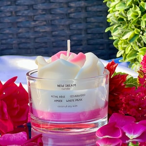 May include: A clear glass candle holder with a white and pink candle shaped like a flower. The candle is labeled "NEW DREAM Candle" and lists scents: Petal Rose, Amber, Cedarwood, and White Musk. The candle is surrounded by red flowers and petals.