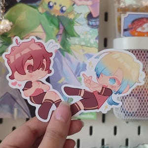 Peut inclure: Deux autocollants de style anime représentant des personnages chibi aux cheveux rouges et bleus, vêtus de tenues marron et rouges. Les autocollants sont tenus dans une main sur un fond d'autres objets liés à l'anime.