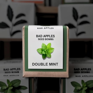 May include: A square package of "Bad Apples Seed Bombs" with a green and white label. The label features the text "BAD APPLES SEED BOMBS" and "DOUBLE MINT" with a sprig of mint. Other packages with similar designs are in the background.