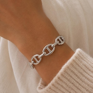 Pode incluir: Uma pulseira de corrente em tom prateado polido, composta por uma série de elos ovais interligados, cada um reforçado com uma barra horizontal central. A pulseira tem um acabamento metálico brilhante e um mecanismo de fecho seguro.