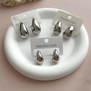 Peut inclure: Trois paires de boucles d'oreilles en forme de goutte d'eau, couleur argent, en acier inoxydable. Chaque paire est fixée sur un carton de présentation blanc rectangulaire portant l'inscription Stainless steel. Finition métallique lisse.
