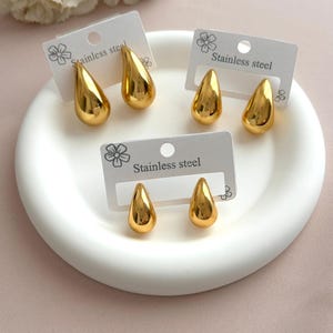 Peut inclure: Trois paires de boucles d'oreilles en forme de goutte, couleur or, en acier inoxydable. Chaque paire est fixée sur une carte de présentation rectangulaire blanche avec une petite icône de fleur et le texte imprimé Stainless steel.