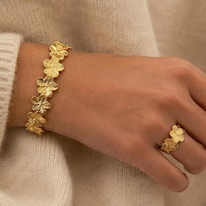 Peut inclure: Un ensemble composé d'un bracelet et d'une bague dorés arborant un motif floral répétitif. Les bijoux présentent une finition mate et texturée, mettant en valeur des formes de fleurs à cinq pétales reliées entre elles pour former les anneaux.
