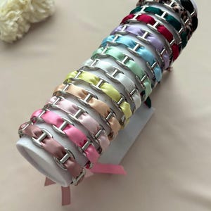 Op de afbeelding: Een collectie haarbanden gerangschikt op een cilindrische display, elk voorzien van een glad satijnen lint in diverse kleuren zoals roze, perzik, geel, wit, mint, blauw, lavendel, rood, donkergroen en luipaardprint met zilverkleurige schakels.