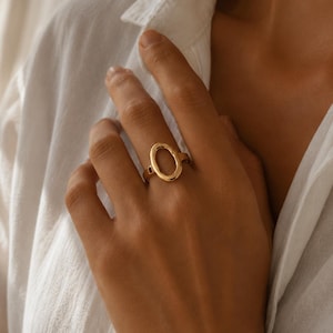 Peut inclure: Une bague en or poli dotée d'un élément central ovale et ouvert. L'anneau est lisse et circulaire, se connectant au cadre ovale vertical. Le métal présente une finition brillante et réfléchissante qui capte la lumière, créant une apparence épurée, simple et structurelle sur le doigt.