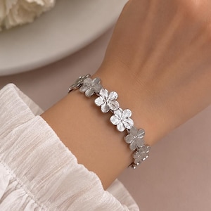 Op de afbeelding: Een zilverkleurige armband bestaande uit een reeks met elkaar verbonden bloemvormige schakels. Elke individuele bloemschakel heeft vijf bloemblaadjes met een getextureerde, geëtste oppervlakteafwerking in een herhalend patroon.