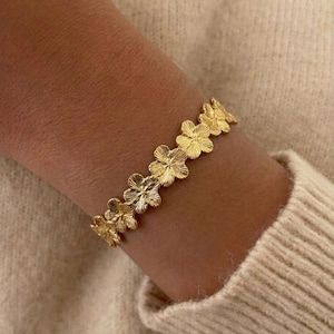 Pode incluir: Uma pulseira de tom dourado com uma série de pingentes de flores de cinco pétalas conectados. Cada flor tem um acabamento de superfície texturizado e radiante que capta a luz. A pulseira é projetada como uma corrente contínua.