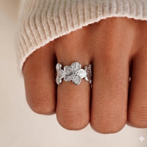 Op de afbeelding: Een zilverkleurige ring met een band die bestaat uit meerdere met elkaar verbonden bloemvormen. Elke bloem heeft gedetailleerde, stralende bloembladtexturen die een dimensionale uitstraling creëren over het gehele oppervlak van de metalen ring.