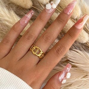 Peut inclure: Une main porte une bague dorée à maillons de chaîne, ornée de longs ongles décorés. Les ongles présentent des motifs variés, dont des paillettes, des perles et des accents floraux en 3D. La bague est le point central, avec un design géométrique audacieux.