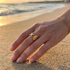 Puede incluir: Un anillo abierto de tono dorado que presenta un motivo de estrella de mar texturizado en un extremo y un diseño de concha marina acanalada en el otro. El anillo se lleva en un dedo y se ve una pulsera de cadena dorada fina y delicada.