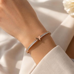 Peut inclure: Un bracelet jonc de couleur or rose orné d'un emblème central en forme de V et d'une rangée de petites pierres claires et scintillantes incrustées le long de l'anneau. Le métal présente une finition polie et un mécanisme d'ouverture à charnière.