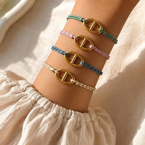 Peut inclure: Quatre bracelets composés de maillons ovales dorés avec une barre horizontale centrale, chacun fixé à un cordon de couleur différente : bleu sarcelle, rose clair, bleu vif et crème. Les bracelets sont portés superposés sur un poignet.