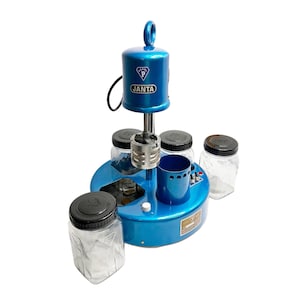 May include: A blue machine with the text "JANTA" on the front. The machine has a circular base with several clear glass jars with black lids. A blue cup and a metal component are also visible.